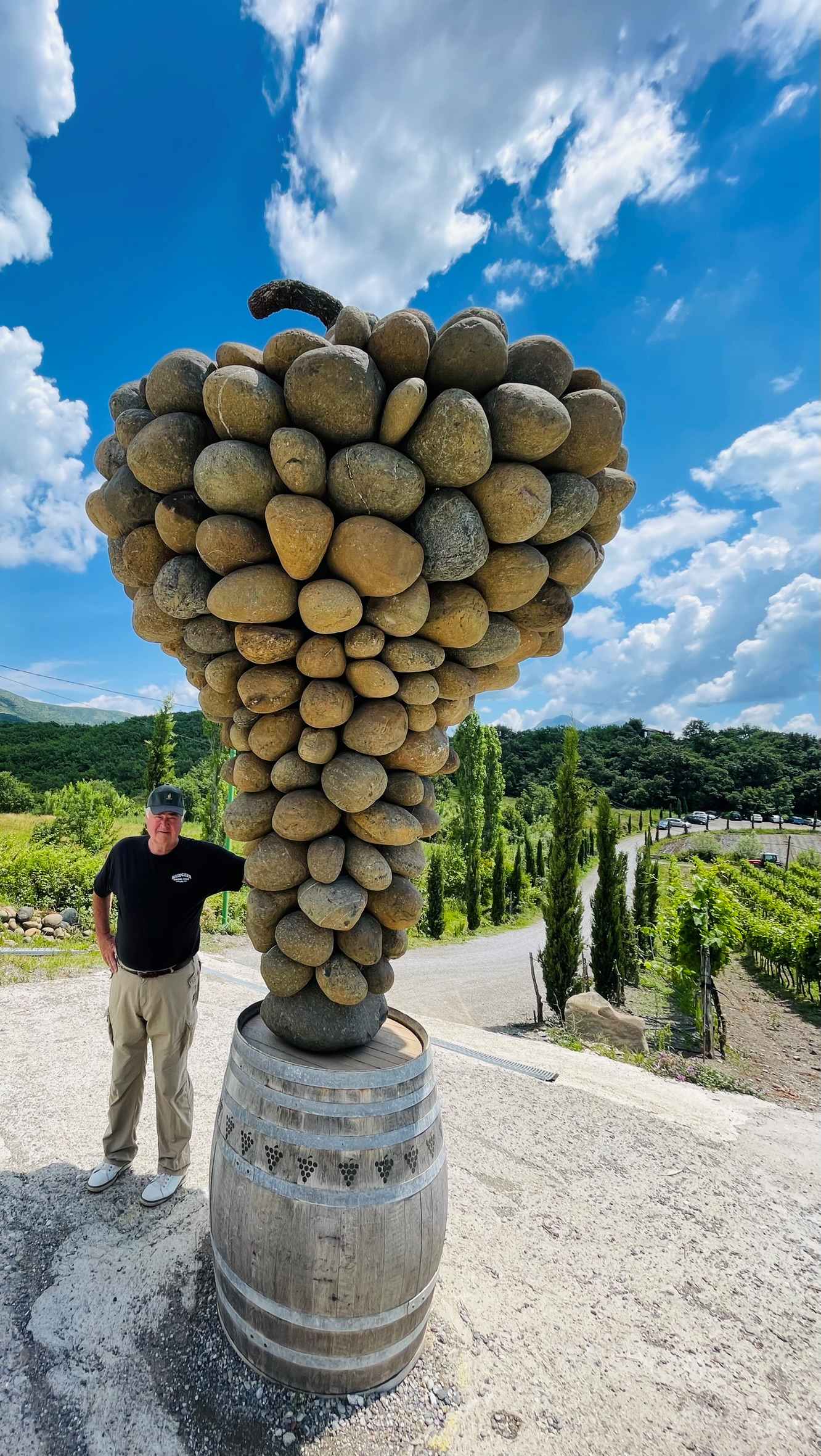 Paul with a sculpture made of large rocks that resemble a grape cluster on a wine barrel in Albania